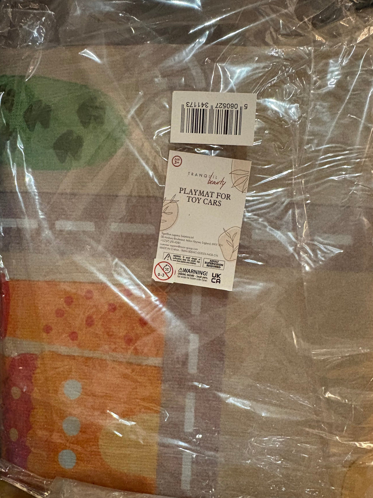 A packaged toy car play mat with a multicolor pattern, wrapped in clear plastic with retail tags attached, sitting on a wooden surface.